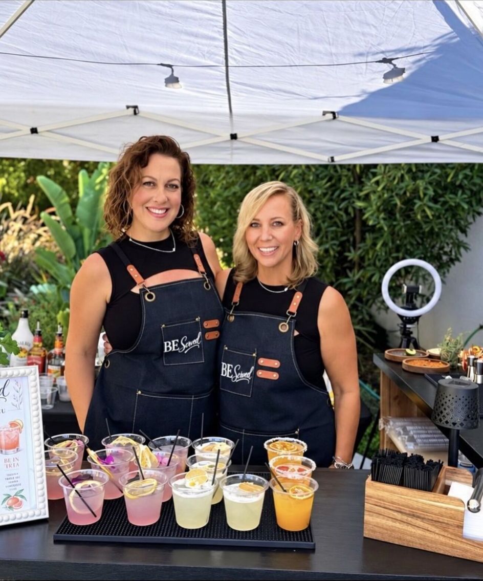 Brooke and Emily, the founders of BeServed, smiling behind their mobile bar at an outdoor event